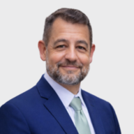 A man with short brown hair and a beard, wearing a navy blue suit, white shirt, and light green tie, smiles at the camera against a plain light background. EFESO Management Consultants